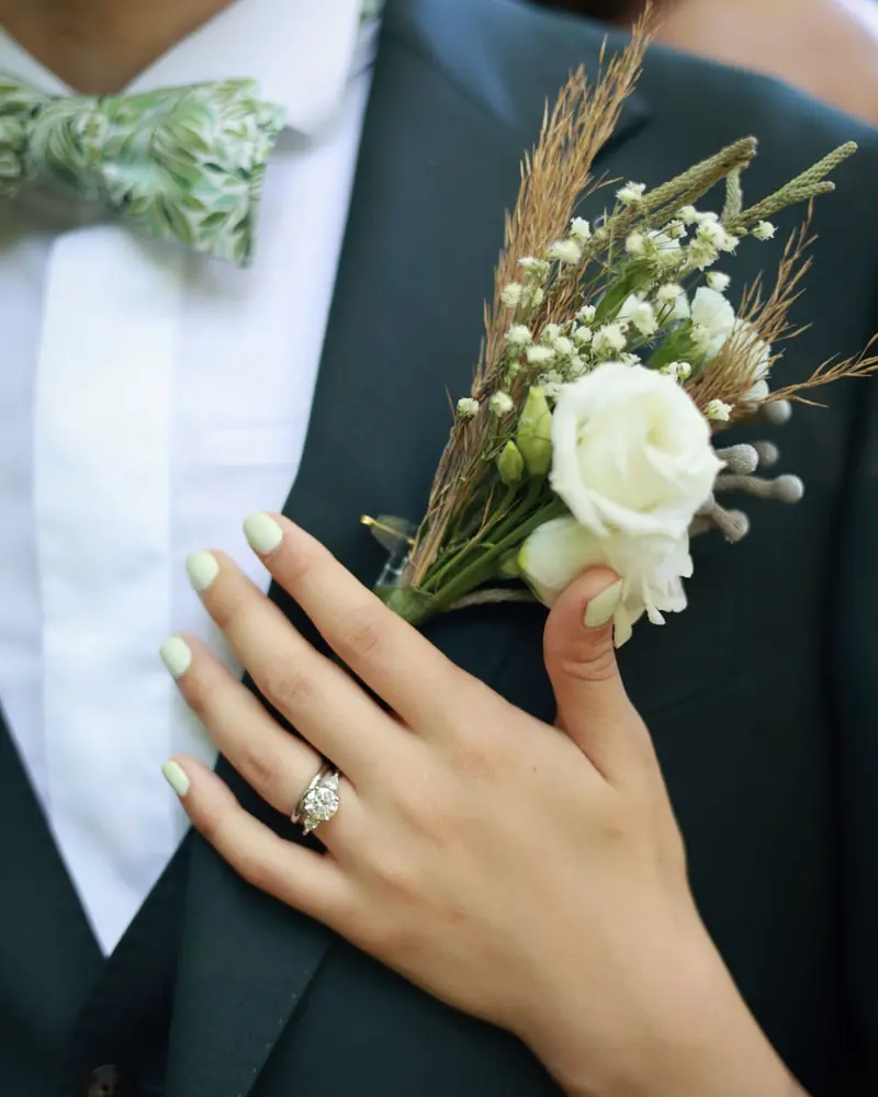 A hand wearing a ring resting on a white flower buttonhole (photo by Vally Photography)