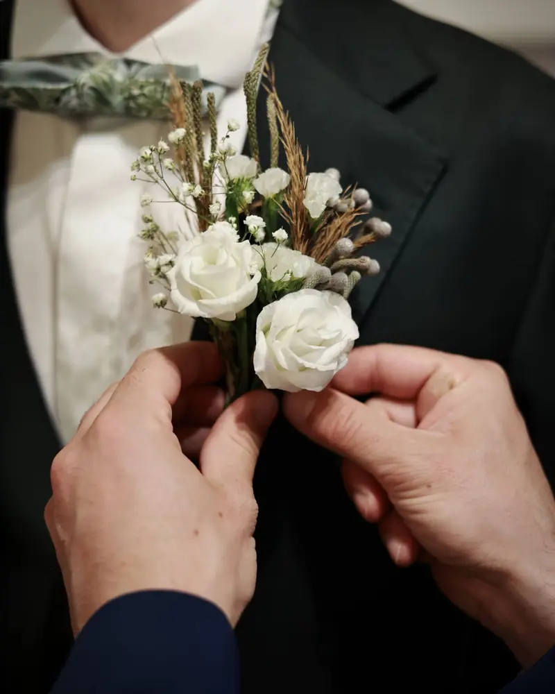 Handen spelden een corsage van witte rozen op een jasje (foto Vally Photography)