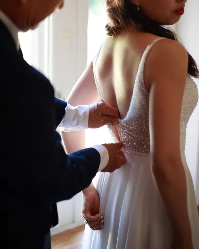 Des mains lacent le dos d'une robe de mariée, juste avant la cérémonie (photo Vally Photography)