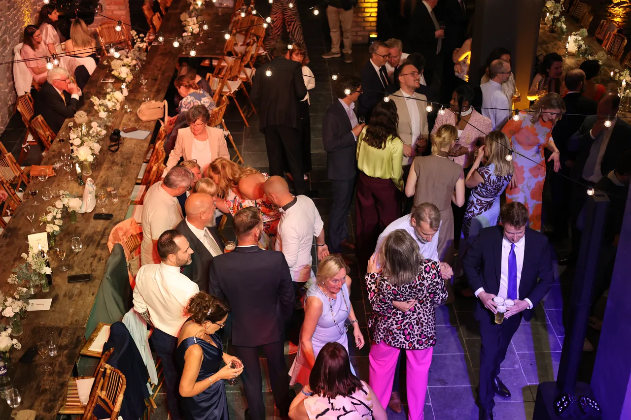 Van bovenaf: de gasten dansen en klinken langs een lange tafel vol bloemen, onder de lichtslingers