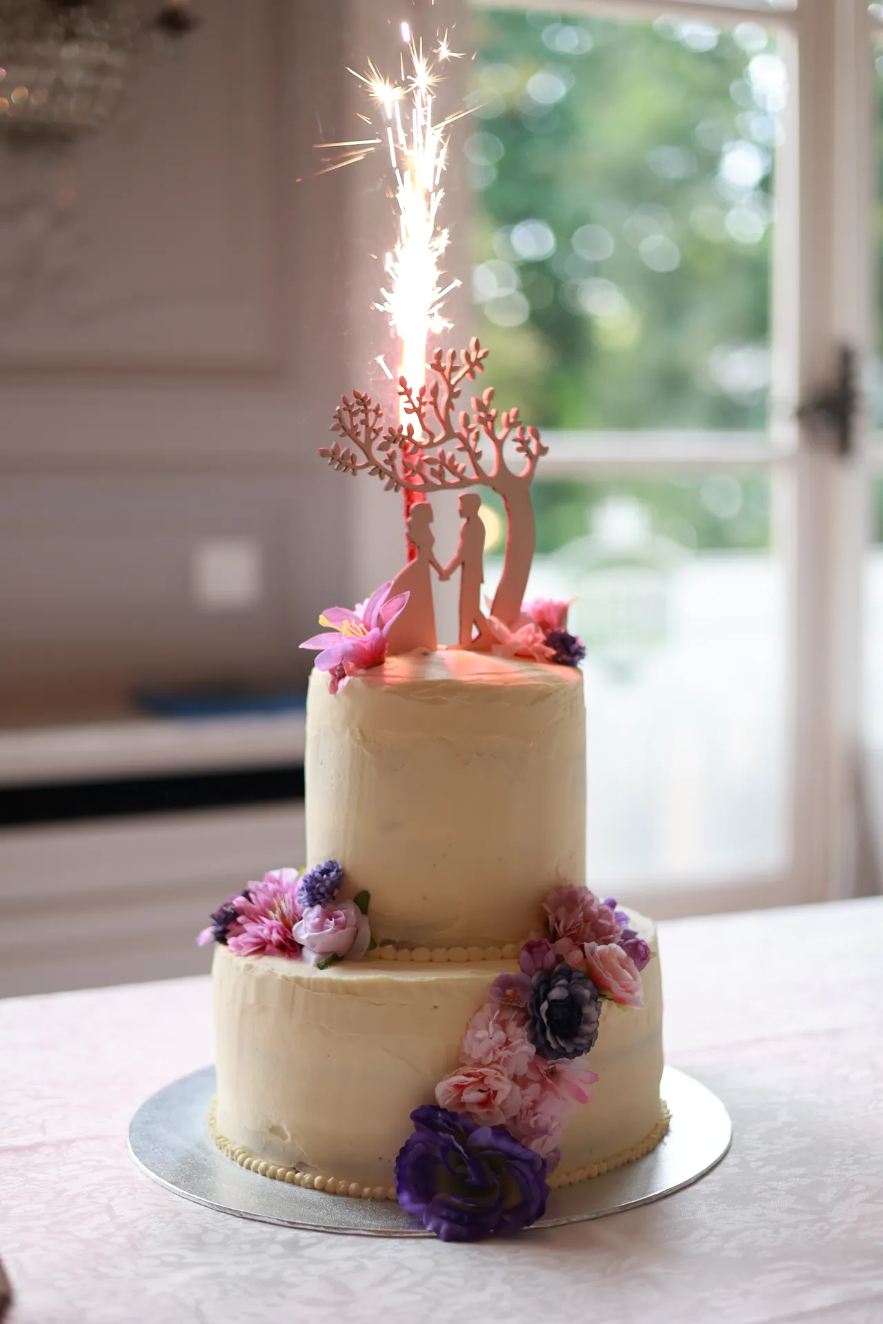 De taart met twee verdiepingen, paarse bloemen en een brandende vonkenfontein