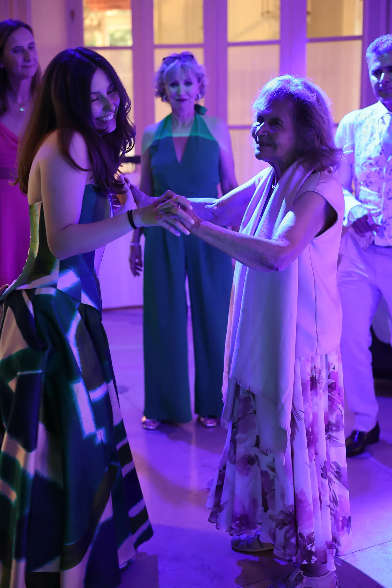 Twee personen dansen hand in hand, de ene lacht, de andere glimlacht, in paars licht