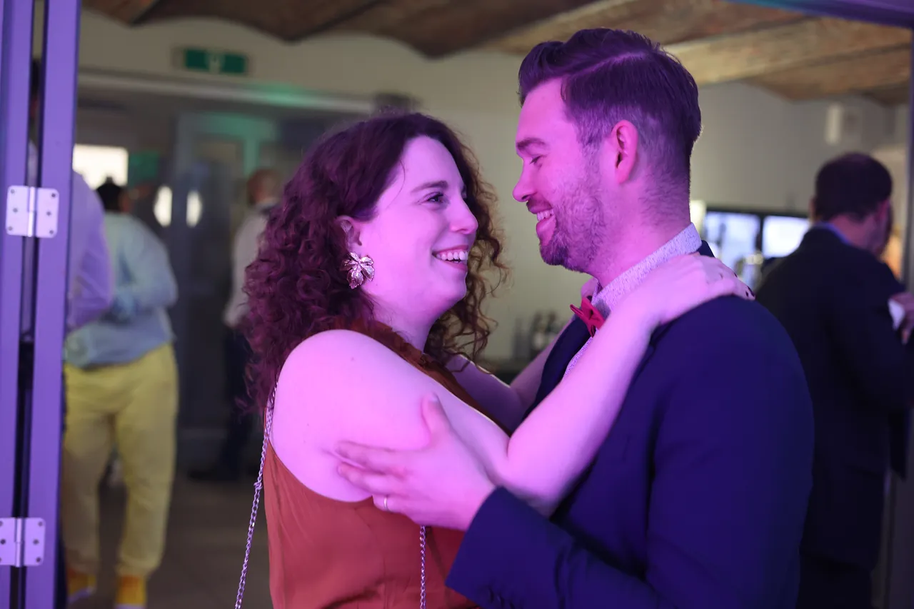 Twee gasten dansen in een omhelzing, gezichten dicht bij elkaar en brede glimlachen, in roze licht