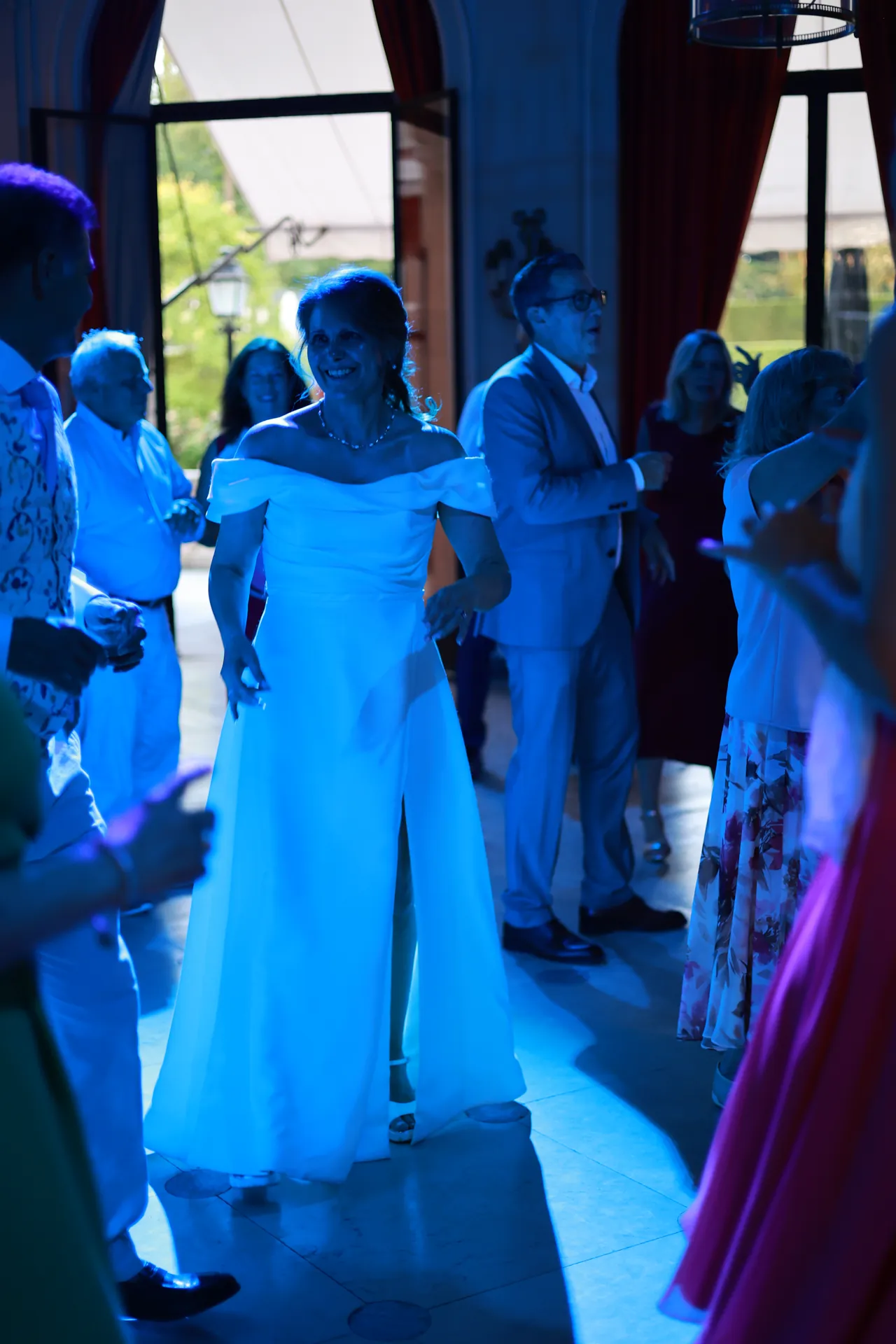 Een persoon in een witte jurk danst glimlachend tussen de gasten, in blauw licht
